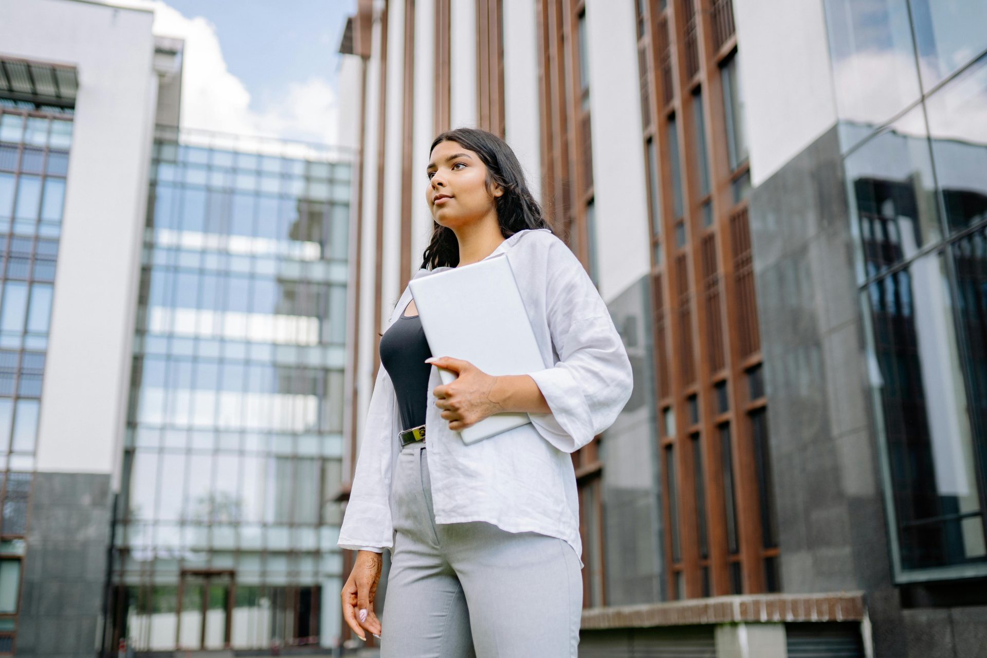 Confident businesswoman stands holding a laptop in front of a modern building.